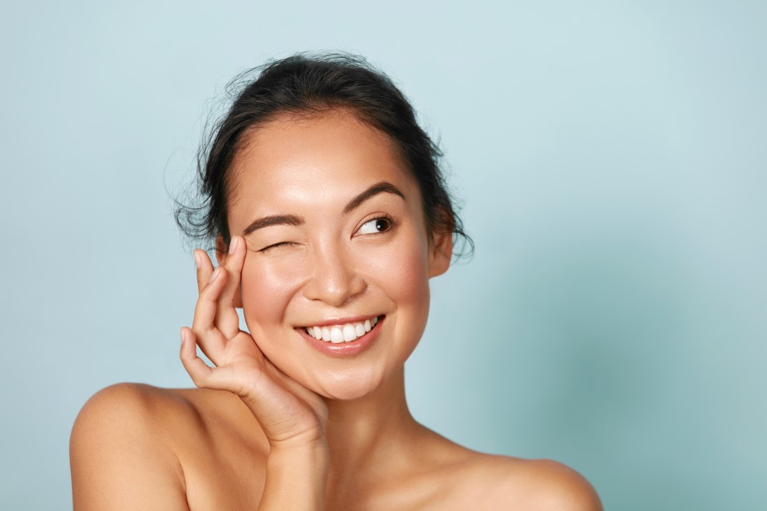 Skin care. Woman with beauty face touching healthy facial skin portrait. Beautiful smiling asian girl model with natural makeup touching glowing hydrated skin on blue background closeup
